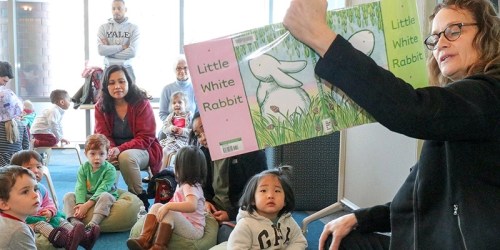 A woman reads "Little White Rabbit" to a group of children sitting on the floor, with adults watching in the background.