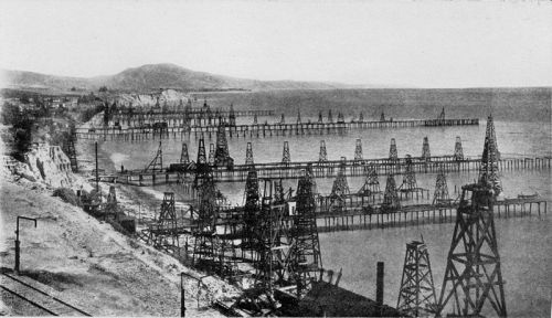 Historic black-and-white photo of oil rigs along a coastline, with mountains in the background and a calm sea.