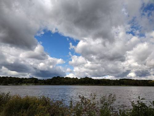 A serene lake scene with a cloudy sky and patches of blue peeking through, surrounded by lush greenery.