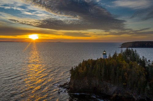 A lighthouse on a rocky shore at sunset, with vibrant clouds and calm waters reflecting the golden light.