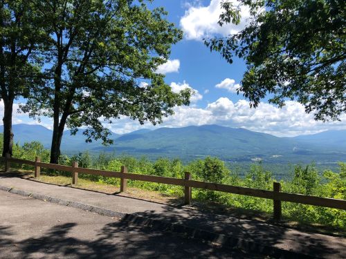 Scenic view of mountains and lush greenery under a blue sky with clouds, framed by trees and a wooden fence.
