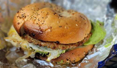 A toasted bagel sandwich filled with sausage, lettuce, and a fried egg, wrapped in foil.