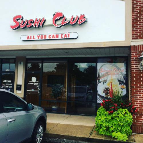 Exterior of Sushi Club restaurant with "All You Can Eat" sign, featuring a car parked outside and vibrant flower planters.