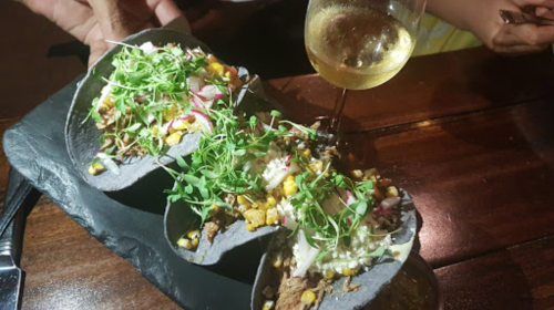 Three black tacos filled with meat, corn, and greens, accompanied by a glass of white wine on a wooden table.