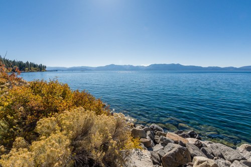 A serene lake view with clear blue water, rocky shore, and colorful foliage under a bright blue sky.