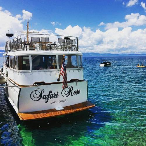 A boat named "Safari Rose" anchored in clear blue water at Lake Tahoe, with a sunny sky and fluffy clouds above.