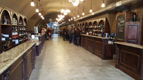 A spacious wine shop with wooden counters, shelves of wine bottles, and customers lined up along the bar.