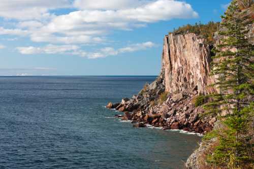 A rocky cliff rises above a calm blue sea under a partly cloudy sky, with trees lining the shore.