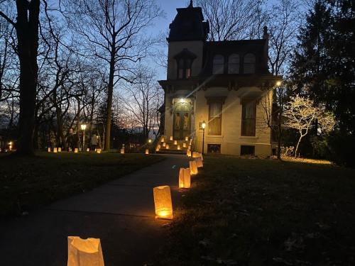 A pathway lined with glowing lanterns leads to a historic house at dusk, surrounded by bare trees.