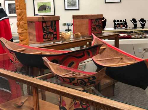 Three intricately designed wooden canoes displayed in a gallery, surrounded by various art pieces and jewelry.