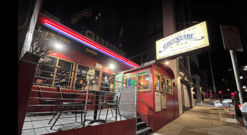A brightly lit diner with a red exterior and neon signs at night, featuring outdoor seating and a welcoming entrance.