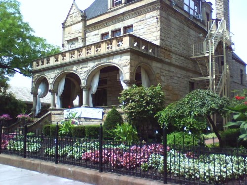 A large stone mansion with arched windows, a balcony, and a lush garden, surrounded by a black fence.