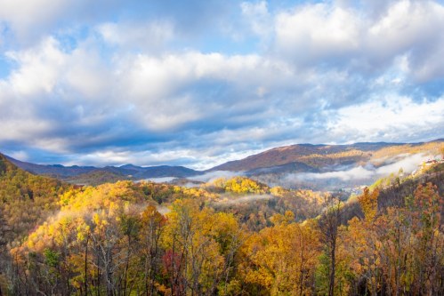 A panoramic view of colorful autumn trees in the mountains, with mist rising and clouds in a blue sky.