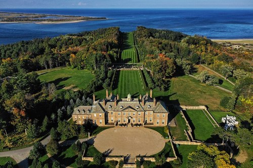 Aerial view of a grand mansion surrounded by lush greenery and a coastline in the background.