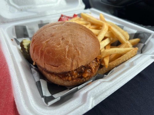 A close-up of a pulled pork sandwich with fries and pickles in a takeout container.