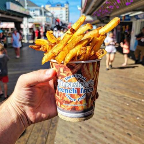 A hand holding a cup of Thrasher's French fries on a boardwalk, with people walking in the background.