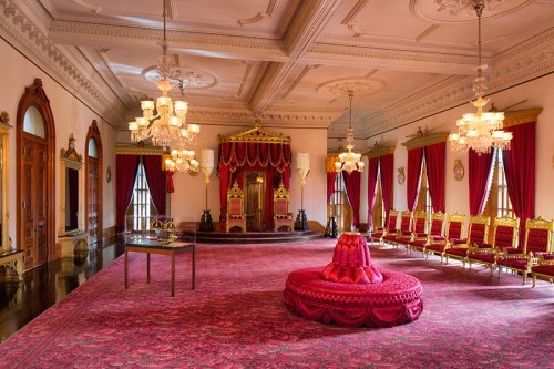 Luxurious room with ornate red decor, a throne, golden chairs, and elegant chandeliers, showcasing opulent design.