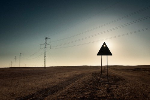 A solitary warning sign stands in a vast, empty landscape with power lines stretching into the horizon under a bright sun.