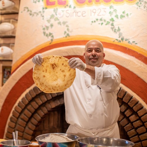 A chef in a white coat holds up a large tortilla in a colorful restaurant setting.