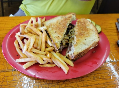 A plate with a sliced sandwich filled with meat and onions, accompanied by a side of golden French fries and pickles.