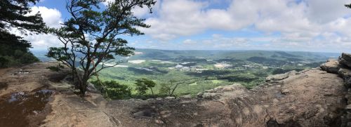 Panoramic view from a rocky overlook, showcasing lush green valleys and distant hills under a partly cloudy sky.