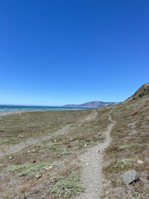 A sandy path leads along the coast, with clear blue skies and distant hills in the background.