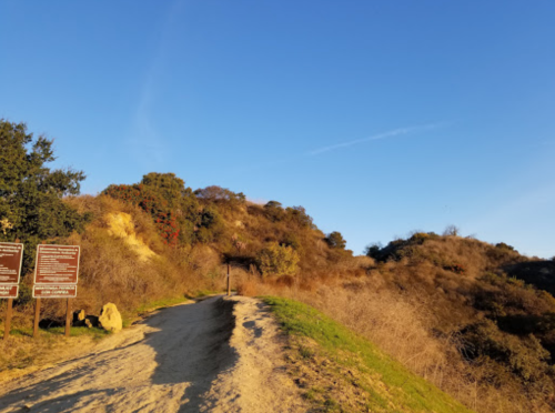 A dirt path leads up a hillside with signs, surrounded by dry vegetation and a clear blue sky.