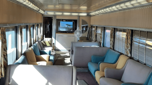 Interior of a vintage train car featuring comfortable seating, large windows, and a fan, with a TV in the background.