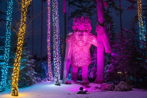 A large wooden troll statue illuminated with pink and yellow lights, surrounded by snowy trees at night.