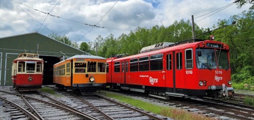 Three vintage trams in red and orange parked on tracks near a green shed, surrounded by trees under a cloudy sky.