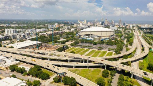 Aerial view of a city skyline with highways, green spaces, and a large stadium under a partly cloudy sky.