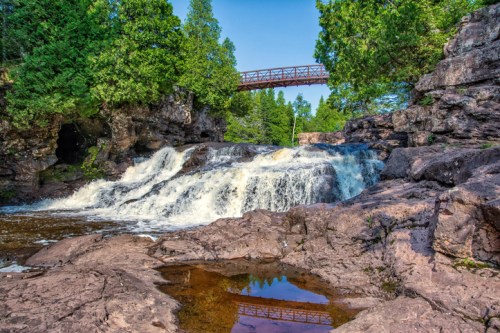 A scenic waterfall cascades over rocky terrain, with a bridge visible above and lush greenery surrounding the area.