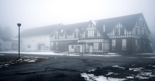 A foggy scene featuring a large, old building with a Tudor-style design, surrounded by snow and a dark, empty parking lot.