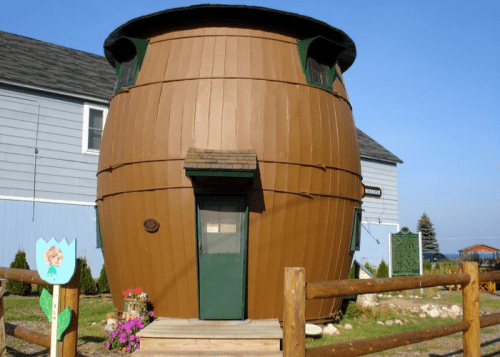 A unique, barrel-shaped house with green windows and a door, surrounded by a wooden fence and flowers.