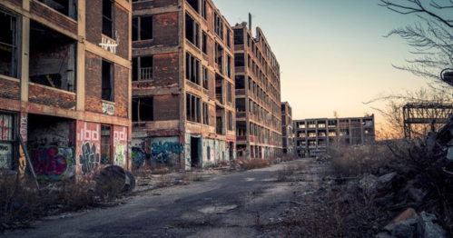 Abandoned industrial buildings with graffiti, overgrown vegetation, and a deserted road at sunset.