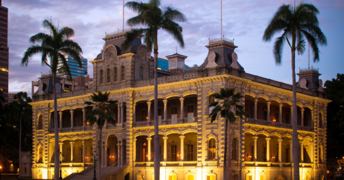 Historic building illuminated at dusk, surrounded by palm trees and city skyline in the background.