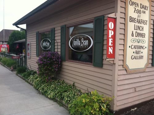 Exterior of Vanilla Bean Restaurant with open sign, flowers, and a sign indicating hours and services offered.