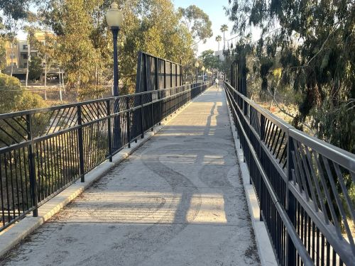 A paved pedestrian bridge surrounded by trees, with shadows cast by the evening sun.