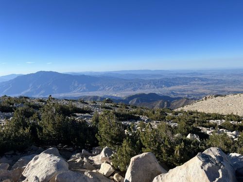 A panoramic view from a mountain peak, showcasing rocky terrain and distant valleys under a clear blue sky.