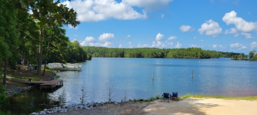 A serene lake surrounded by trees, with a sandy shore and a dock extending into the water under a blue sky.