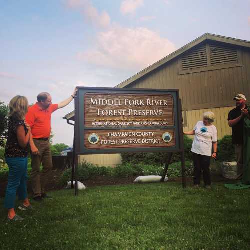 Group of people gathered around a sign for Middle Fork River Forest Preserve, with a building and blue sky in the background.