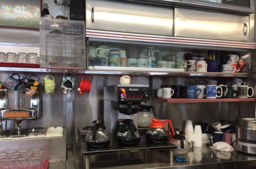 A coffee station with a Bunn coffee maker, various mugs on shelves, and colorful decorations hanging nearby.