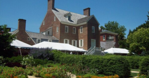 A large brick house with a slate roof, surrounded by lush greenery and a garden, under a clear blue sky.