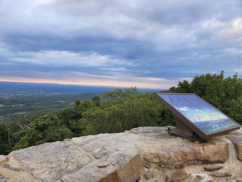 A scenic view from a rocky overlook, featuring a viewing panel and a cloudy sky at sunset.