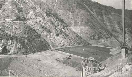 Black and white image of a mountainous landscape with a construction site and machinery in a valley below.