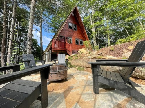 A cozy A-frame cabin surrounded by trees, with a stone patio and black chairs near a fire pit.
