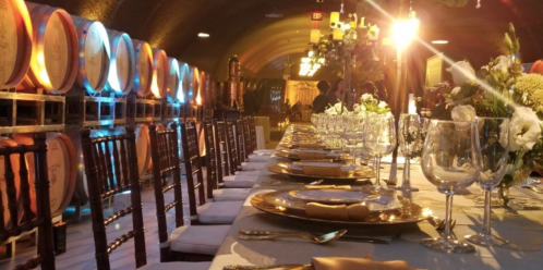 Elegant dining setup in a wine cellar, featuring a long table with fine china and wine barrels in the background.