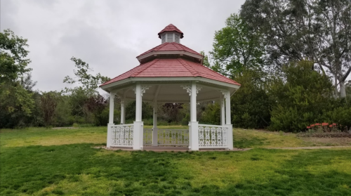 A white gazebo with a red roof stands in a grassy area surrounded by trees and shrubs under a cloudy sky.