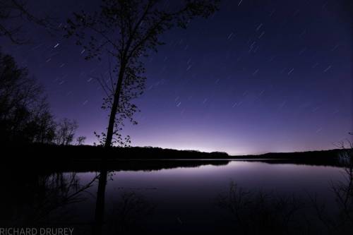 A serene night scene of a calm lake reflecting the starry sky, framed by silhouetted trees along the shore.