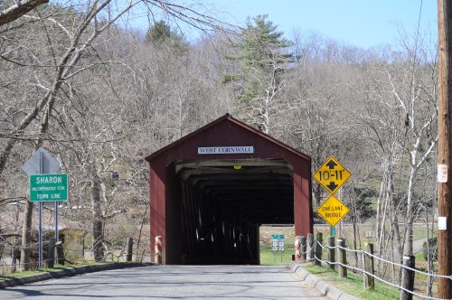 A red covered bridge with a sign reading "West Cornwall," surrounded by bare trees and a clear blue sky.